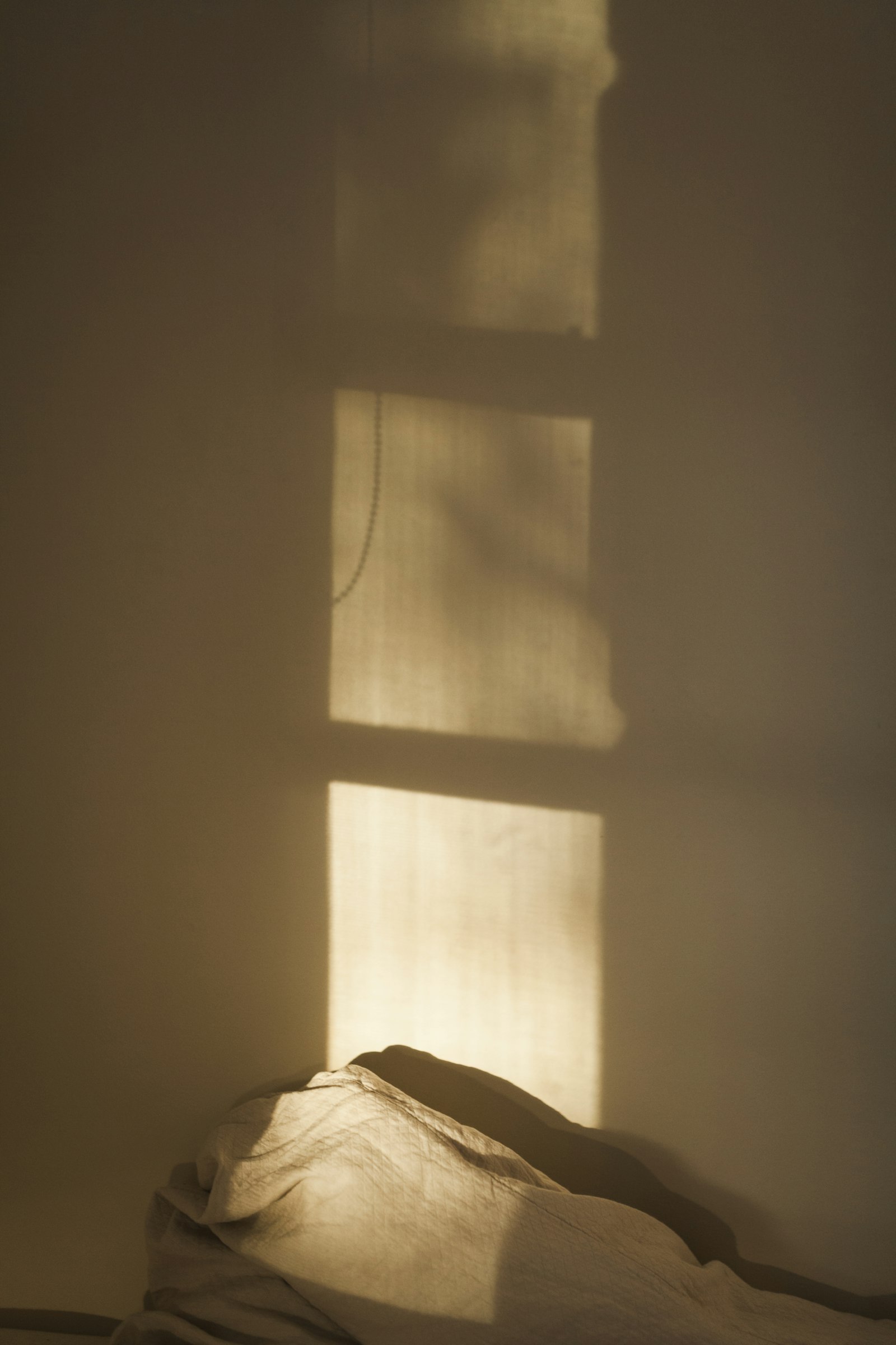 Morning sun casting the shadow of a window across a wall and unmade linen.